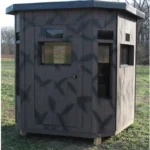 County line dark brown wooden hunting blind with door and windows in hunting field.