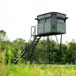 Redneck elevated hunting blind with dark green exterior located on a field.