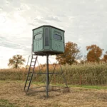Redneck elevated hunting blind with dark green exterior located on a field.