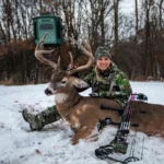 Female hunter wearing all camouflage with a deer that she hunted and a redneck hunting blind in the background.