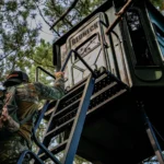 Redneck hunting blinds amongst trees with a hunter wearing camouflage climbing up the ladder.