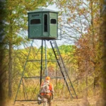 Redneck elevated hunting blind with dark green exterior in the woods. Hunter with camouflage and gun walking towards blind.