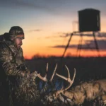 Male hunter in camouflage sitting with dead deer that he hunted, hunting blind behind him.
