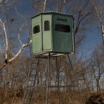 Redneck elevated hunting blind with dark green exterior located in woods.
