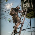Redneck hunting blind with hunter carrying bow in hand wearing all camouflage climbing up hunting blind ladder.